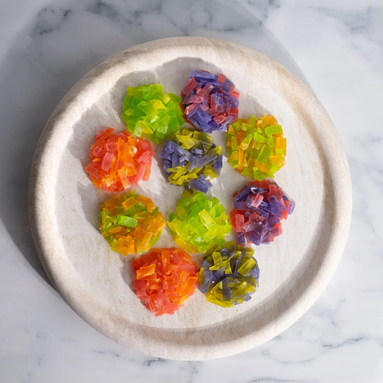 Overhead of ten multi-colour crystal candy clusters arranged on a round marble table; thin-slice clusters with ASMR crunch.