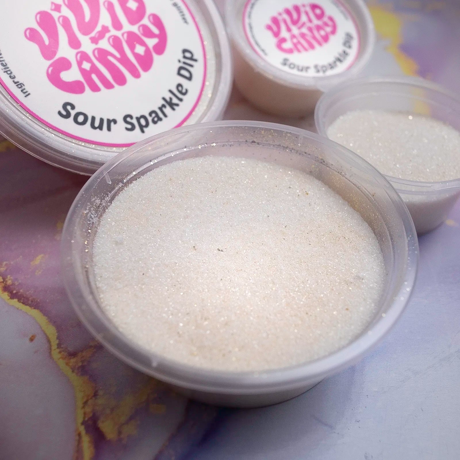 Close view of an open tub showing the glittery sour sugar texture of Sour Sparkle Dip; branded lids in the background.