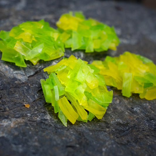 Close-up of lime and pineapple crystal candy clusters on dark rock; neon green/yellow thin-cut shards with jewel-like shine.