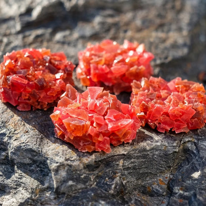 Four clusters of bright red-brown crystal candy clusters placed on a dark textured rock surface.