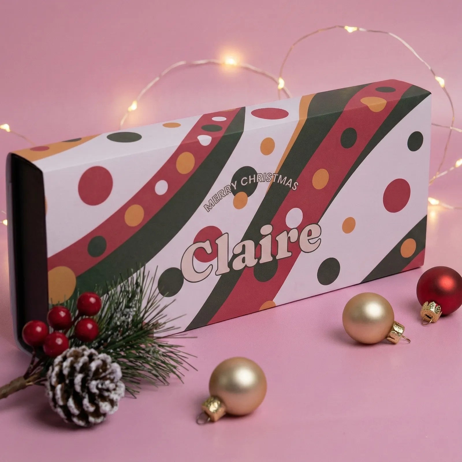 Decorative Christmas gift box with polka dots and text, surrounded by festive ornaments on a pink background.  Custom printed name
