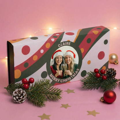Personalized Christmas gift box with polka dot design and photo of two people, surrounded by festive decorations on a pink background.