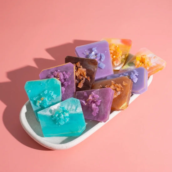 A group of colorful rectangular handmade crystal candy of turquoise, purple, brown, and orange, each topped with dried flower petals, arranged on a white ceramic dish against a solid pink background.