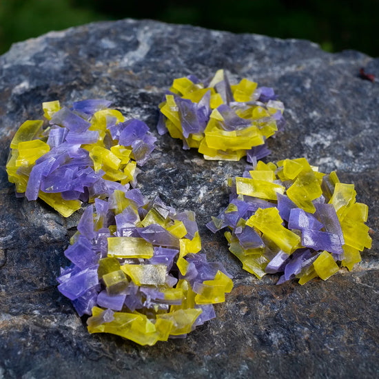 Purple and yellow thin-slice crystal candy clusters (pineapple + blueberry) clustered on dark rock; glossy shards with crunchy snap.