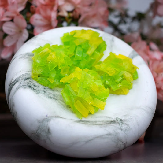 Lime-green and yellow crystal candy clusters arranged on a marble plate in front of pink flowers; thin-slice design for clean crunch.