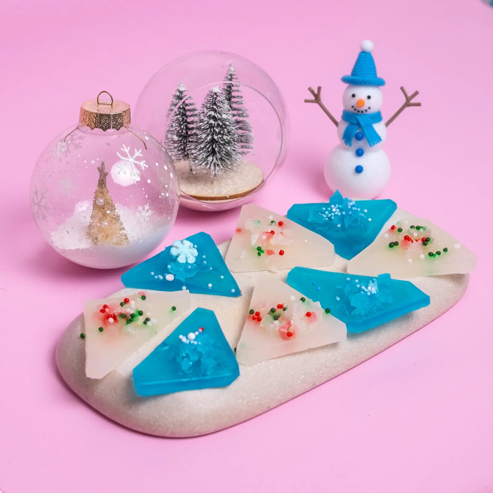 A Blue and white Japanese style crystal candy with a snow globe, miniature trees, and a snowman on a pink background.