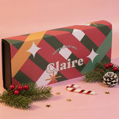 Christmas-themed custom printed gift box with geometric pattern and 'Merry Christmas Claire' text, surrounded by festive decorations on a pink background.